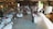 A rustic venue is decorated for a wedding reception featuring round tables with white tablecloths, floral centerpieces, and guests seated around them. A couple is dancing in the center under soft lighting. String lights and draped fabric add to the cozy atmosphere. On the sides, people are observing the dance, and there's a vintage ambiance with wooden walls and soft green accents.