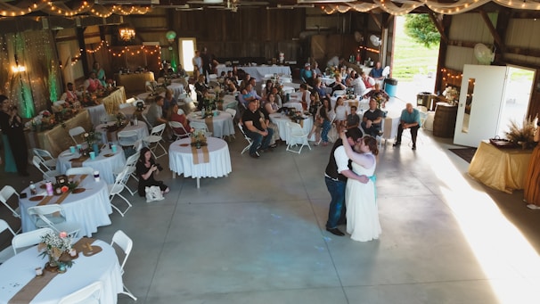 A rustic venue is decorated for a wedding reception featuring round tables with white tablecloths, floral centerpieces, and guests seated around them. A couple is dancing in the center under soft lighting. String lights and draped fabric add to the cozy atmosphere. On the sides, people are observing the dance, and there's a vintage ambiance with wooden walls and soft green accents.
