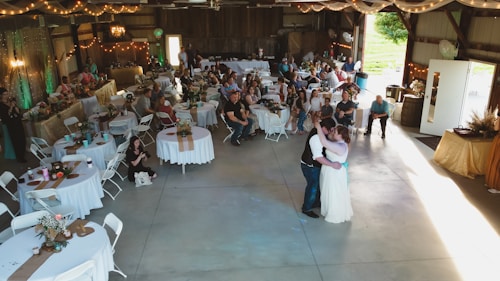 A rustic venue is decorated for a wedding reception featuring round tables with white tablecloths, floral centerpieces, and guests seated around them. A couple is dancing in the center under soft lighting. String lights and draped fabric add to the cozy atmosphere. On the sides, people are observing the dance, and there's a vintage ambiance with wooden walls and soft green accents.