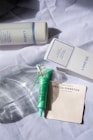 A collection of skincare products is arranged on a crumpled white fabric. Visible items include a Laneige Cream Skin toner and its box, a green skincare tube, a clear glass bottle, and a pack of Mecca Cosmetica blotting papers.