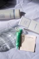 A collection of skincare products is arranged on a crumpled white fabric. Visible items include a Laneige Cream Skin toner and its box, a green skincare tube, a clear glass bottle, and a pack of Mecca Cosmetica blotting papers.