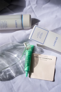 A collection of skincare products is arranged on a crumpled white fabric. Visible items include a Laneige Cream Skin toner and its box, a green skincare tube, a clear glass bottle, and a pack of Mecca Cosmetica blotting papers.