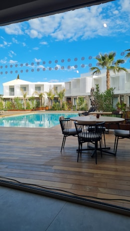 A modern outdoor patio area looking out onto a swimming pool, surrounded by contemporary white buildings and palm trees. The sky is clear with scattered clouds, and there is a set of patio furniture including a round table and chairs on a wooden deck.