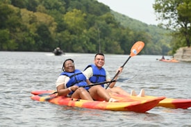 Two people are joyfully kayaking on a river, both wearing blue life vests. They are paddling in tandem on a tandem kayak, surrounded by lush green trees along the riverbank. The water ripples around them as other kayakers can be seen in the background.