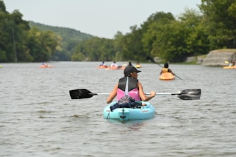 A group of people kayaking on a calm river surrounded by lush green trees. In the foreground, a person wearing a life jacket and hat is paddling a blue kayak, with several other kayakers visible in the distance. The setting is serene and peaceful.
