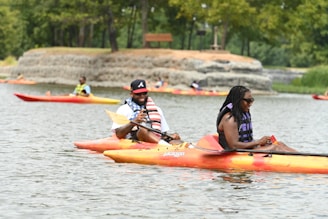 Two people are leisurely kayaking on a calm lake. They are smiling and appear relaxed. Around them, other kayakers can be seen enjoying the water. Trees and a small rock formation are visible in the background, under a clear sky.