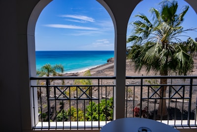A scenic view of a tropical beach seen through arched windows on a balcony. The ocean is a vivid blue, meeting a clear sky, with a sandy beach at the shoreline. Palm trees and lush greenery are visible on the foreground and the sides, providing a contrast to the rocky terrain.