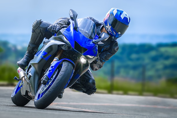 A motorcyclist wearing a blue helmet and full protective gear leans sharply into a turn on a sleek blue sport bike. The background is blurred, suggesting high speed, with green landscape visible, indicating an outdoor setting.