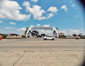 An ornate archway spans across a road with a white car and a motorbike passing by. The archway has signage indicating Surat Airport, with trees and greenery flanking either side. The sky is clear with scattered clouds, creating a bright and open atmosphere.