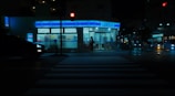 A convenience store with bright interior lighting is located at a street intersection during nighttime. The crosswalk in the foreground is dimly lit, and a silhouette of a person is visible approaching the store. A car with its headlights on is passing by on the left side.