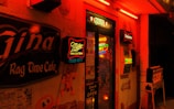 A dimly lit bar entrance adorned with neon signs advertising brands like Miller High Life and Budweiser. The exterior is illuminated in warm red and orange hues, with vintage-style decorations and signage indicating it is a café and bar. Posters and a 'Come in We're Open' sign enhance the retro vibe.