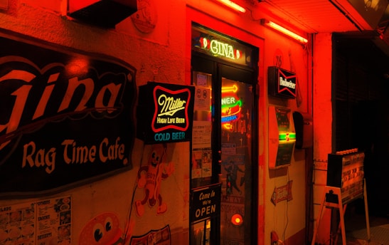 A dimly lit bar entrance adorned with neon signs advertising brands like Miller High Life and Budweiser. The exterior is illuminated in warm red and orange hues, with vintage-style decorations and signage indicating it is a café and bar. Posters and a 'Come in We're Open' sign enhance the retro vibe.