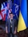 A person is standing in front of a row of international flags. The individual is wearing a blue blazer over a dark shirt and has a lanyard around their neck. The flags behind the person include those from the United States, the United Kingdom, and other nations, as well as some organizational flags. The setting appears to be indoors with a patterned carpet visible on the floor.