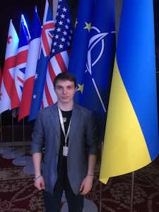 A person is standing in front of a row of international flags. The individual is wearing a blue blazer over a dark shirt and has a lanyard around their neck. The flags behind the person include those from the United States, the United Kingdom, and other nations, as well as some organizational flags. The setting appears to be indoors with a patterned carpet visible on the floor.