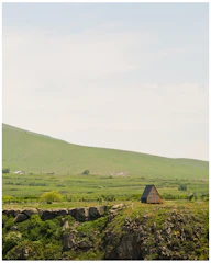 A small A-frame cabin is situated on a rocky cliff surrounded by lush greenery and rolling hills. The cabin's dark roof contrasts with its wooden structure. The landscape is expansive, with a clear sky overhead and a vast green field stretching into the distance.