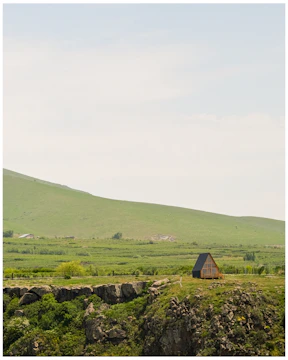 A small A-frame cabin is situated on a rocky cliff surrounded by lush greenery and rolling hills. The cabin's dark roof contrasts with its wooden structure. The landscape is expansive, with a clear sky overhead and a vast green field stretching into the distance.