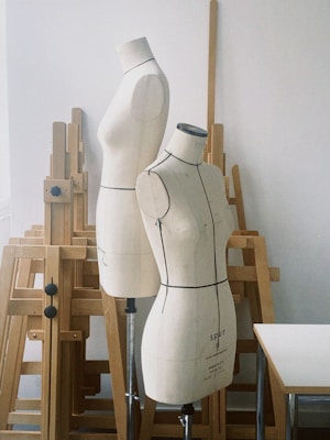 Two dressmaker's mannequins are standing among several wooden easels in an artist's or designer's studio. The mannequins have a minimalist design with clear outlines for garment construction. The background includes various wooden easels and a white table, suggesting a creative workspace.