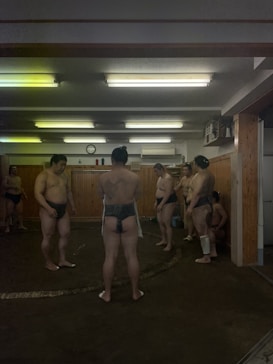 In an indoor training room with wooden walls, several sumo wrestlers are gathered, dressed in traditional mawashi belts. A faintly illuminated space with fluorescent lighting overhead creates a focused yet subdued atmosphere. The wrestlers stand on a circular ring made of distinct material, engaging in what appears to be training or practice.