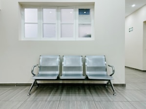 A public waiting area featuring a row of three metallic chairs placed against a pale wall. Above the chairs is a set of frosted windows, and the flooring is made of gray tiles. The space appears clean, minimalist, and well-maintained, often typical of a hospital or office corridor.