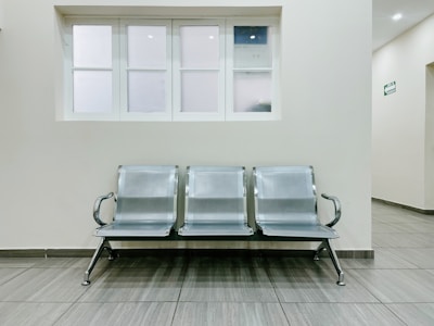 A public waiting area featuring a row of three metallic chairs placed against a pale wall. Above the chairs is a set of frosted windows, and the flooring is made of gray tiles. The space appears clean, minimalist, and well-maintained, often typical of a hospital or office corridor.