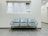 A public waiting area featuring a row of three metallic chairs placed against a pale wall. Above the chairs is a set of frosted windows, and the flooring is made of gray tiles. The space appears clean, minimalist, and well-maintained, often typical of a hospital or office corridor.
