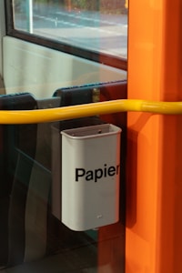 A grey paper bin with the word 'Papier' printed on it is mounted on a large window with a bright yellow handlebar in front. The setting appears to be inside a vehicle, possibly a train or bus, with an orange vertical panel adjacent to the bin.