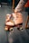 A pair of vintage-style roller skates is being held by a person. The skates are light pink with laces and studs, and they have a colorful rainbow strap. The setting appears to be indoors, possibly on a bus or subway, indicated by the pole and seat in the background.