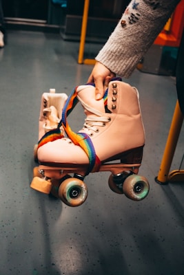 A pair of vintage-style roller skates is being held by a person. The skates are light pink with laces and studs, and they have a colorful rainbow strap. The setting appears to be indoors, possibly on a bus or subway, indicated by the pole and seat in the background.