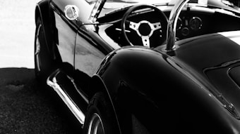 A sleek, black convertible car with a shiny finish is parked, showcasing its smooth curves and classic design. The image focuses on the rear and side view, highlighting the open-top aspect and the stylish steering wheel inside.