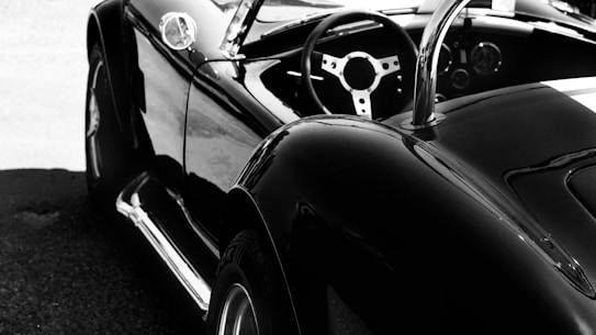 A sleek, black convertible car with a shiny finish is parked, showcasing its smooth curves and classic design. The image focuses on the rear and side view, highlighting the open-top aspect and the stylish steering wheel inside.