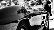 A sleek, reflective surface of a classic car is prominently displayed with detailed contours. The shiny exterior captures reflections of nearby trees and the environment. In the background, blurred figures of people are visible standing in a sunny outdoor setting.