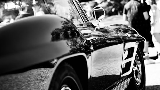 A sleek, reflective surface of a classic car is prominently displayed with detailed contours. The shiny exterior captures reflections of nearby trees and the environment. In the background, blurred figures of people are visible standing in a sunny outdoor setting.