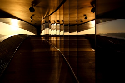 A modern architectural space with reflective glass walls on one side, capturing the warmth of the setting sun. The play of light and shadows creates a sense of depth and movement. The overall structure appears sleek and contemporary.