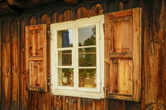 A rustic wooden window with shutters on both sides is set into a wooden cabin wall. The weathered wood has a warm, natural texture, reflecting a golden hue. Inside, vintage bottles can be seen through the clear glass, placed on a shelf. A partial reflection of the surrounding greenery is visible on the window pane.
