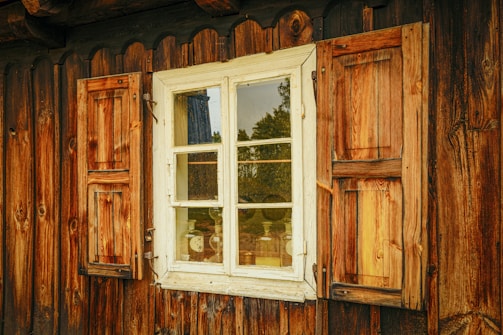 A rustic wooden window with shutters on both sides is set into a wooden cabin wall. The weathered wood has a warm, natural texture, reflecting a golden hue. Inside, vintage bottles can be seen through the clear glass, placed on a shelf. A partial reflection of the surrounding greenery is visible on the window pane.
