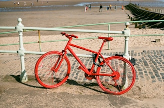 A bright red bicycle leans against a mint green railing near a sandy beach. The surrounding area includes a cobblestone path and a few people in the distance enjoying the beach environment and the sea.