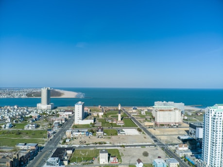 A coastal urban landscape featuring several multi-story buildings with a vast body of water in the background. The shoreline curves gently, with a sandy beach extending into the blue sea. Streets are visible with a grid-like layout, surrounded by various residential and commercial structures.