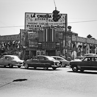 A busy urban intersection featuring vintage cars from the mid-20th century, with a large sign for 'La Ciguena Club' in the background. The scene depicts a lively street with multiple parked cars and people in motion. The architecture and advertisements suggest a vibrant nightlife area.