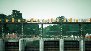 An industrial structure with a series of gates and railings is positioned over a body of water, surrounded by lush green trees. The text 'KANJLI X REGULATOR RD 207730 HOLY BEIN' is displayed on a sign. The structure has a series of orange and yellow markings, contributing to its utilitarian appearance.