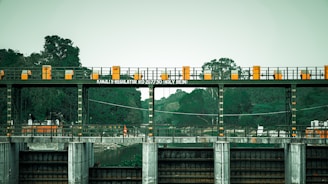 An industrial structure with a series of gates and railings is positioned over a body of water, surrounded by lush green trees. The text 'KANJLI X REGULATOR RD 207730 HOLY BEIN' is displayed on a sign. The structure has a series of orange and yellow markings, contributing to its utilitarian appearance.