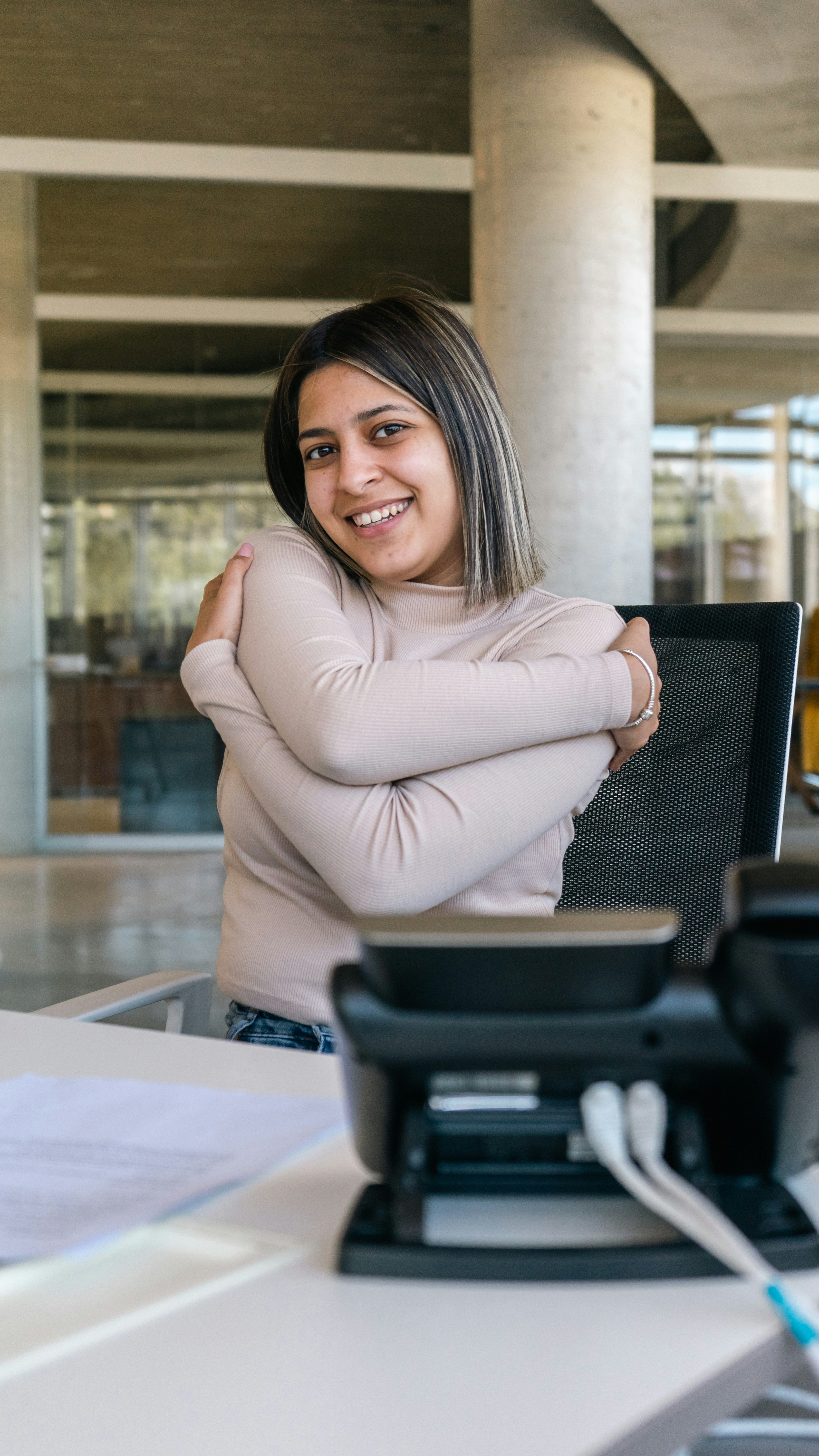Mujer sonriente con blusa beige sentada en un escritorio.
