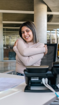 A person sitting in an office environment, smiling, with arms crossed in a self-hugging gesture. A desk phone and some papers are visible on the table in the foreground. The setting includes modern architecture with glass and concrete elements.