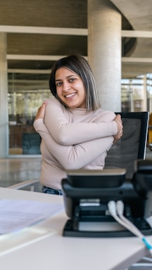 A person sitting in an office environment, smiling, with arms crossed in a self-hugging gesture. A desk phone and some papers are visible on the table in the foreground. The setting includes modern architecture with glass and concrete elements.