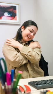 A person with long dark hair is seated at a desk, embracing themselves with a gentle smile. In front of them on the desk are various office supplies including pens and markers. A framed picture of an eye is visible on the wall in the background.
