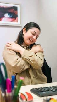 A person with long dark hair is seated at a desk, embracing themselves with a gentle smile. In front of them on the desk are various office supplies including pens and markers. A framed picture of an eye is visible on the wall in the background.