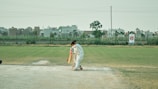 A cricketer wearing white protective gear is batting on a grassy field. In the background, there are buildings and a clear sky. The batsman appears to be focused on playing a shot, positioned near a cricket pitch.