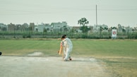 A cricketer wearing white protective gear is batting on a grassy field. In the background, there are buildings and a clear sky. The batsman appears to be focused on playing a shot, positioned near a cricket pitch.