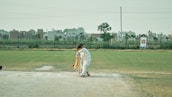 A cricketer wearing white protective gear is batting on a grassy field. In the background, there are buildings and a clear sky. The batsman appears to be focused on playing a shot, positioned near a cricket pitch.
