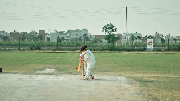 A cricketer wearing white protective gear is batting on a grassy field. In the background, there are buildings and a clear sky. The batsman appears to be focused on playing a shot, positioned near a cricket pitch.