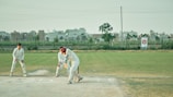 The image captures a group of people engaged in a cricket match. A batsman, wearing a white uniform, is in the act of playing a shot. Behind the batsman, a wicketkeeper and a fielder are positioned, also in white uniforms. The cricket field is green with a boundary line in the background. Beyond the field, there are buildings and a tree, with a banner visible on the right side.